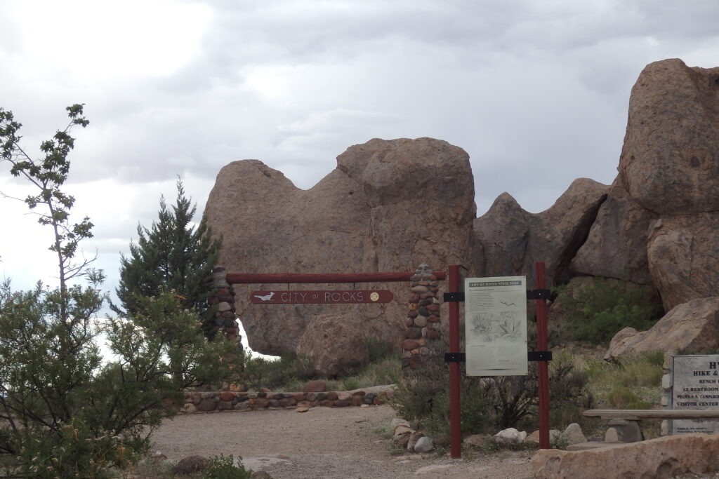 Entrance sign for City of Rocks State Park, framed by large rock formations, with an informational panel nearby outlining park features.