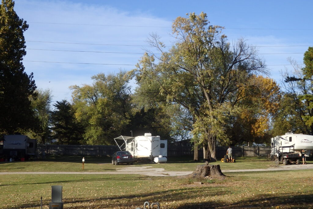 Two RVs are parked in a grassy area surrounded by trees, with one RV's awning extended. A person operates garden equipment nearby.