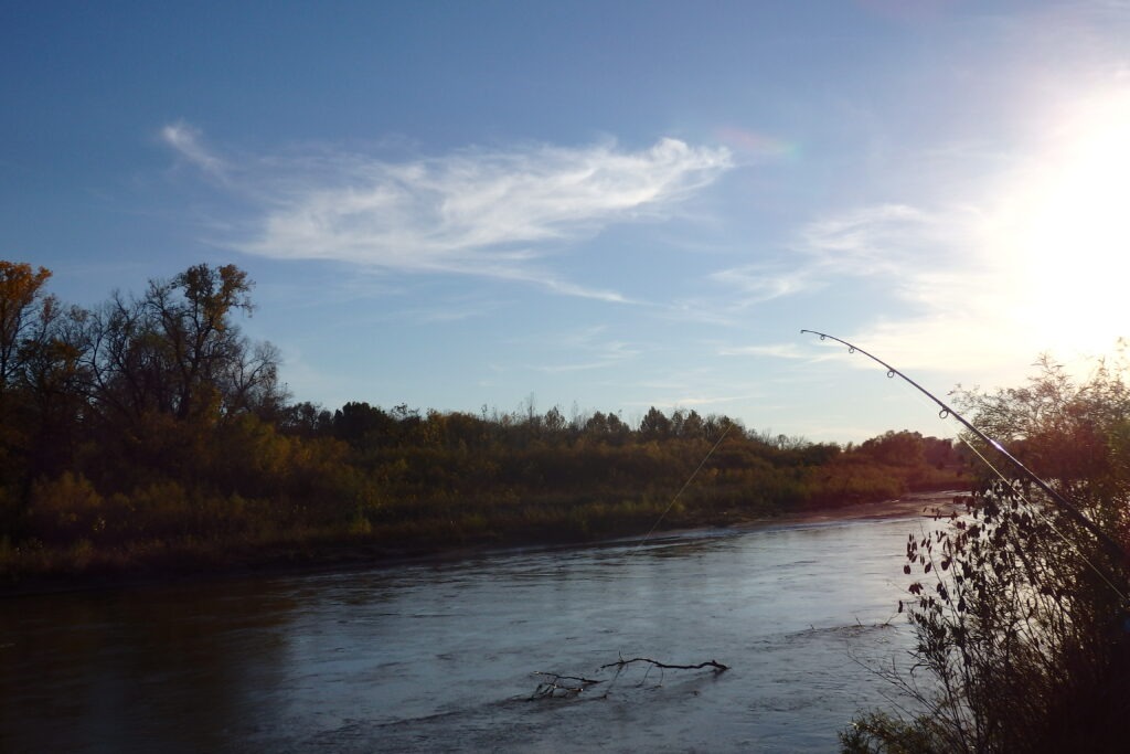 Autumn landscape by a tranquil river, featuring a fishing rod leaning over the water, under a clear blue sky. Perfect for outdoor enthusiasts.