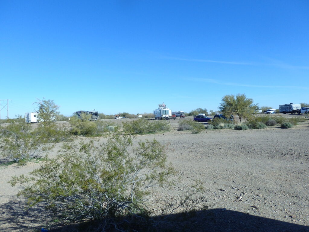 RV park scene in a desert landscape, showcasing several parked recreational vehicles among sparse vegetation under a clear blue sky.