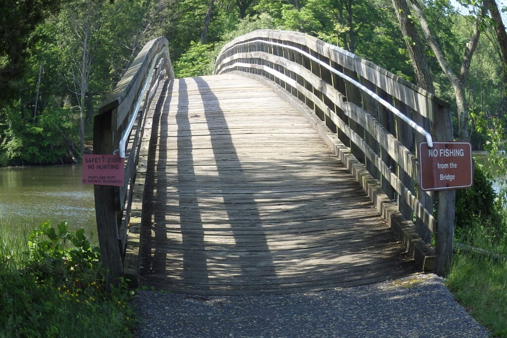 Wooden bridge over a calm river, flanked by greenery. Signs indicate a safety zone with no hunting or fishing allowed from the bridge.