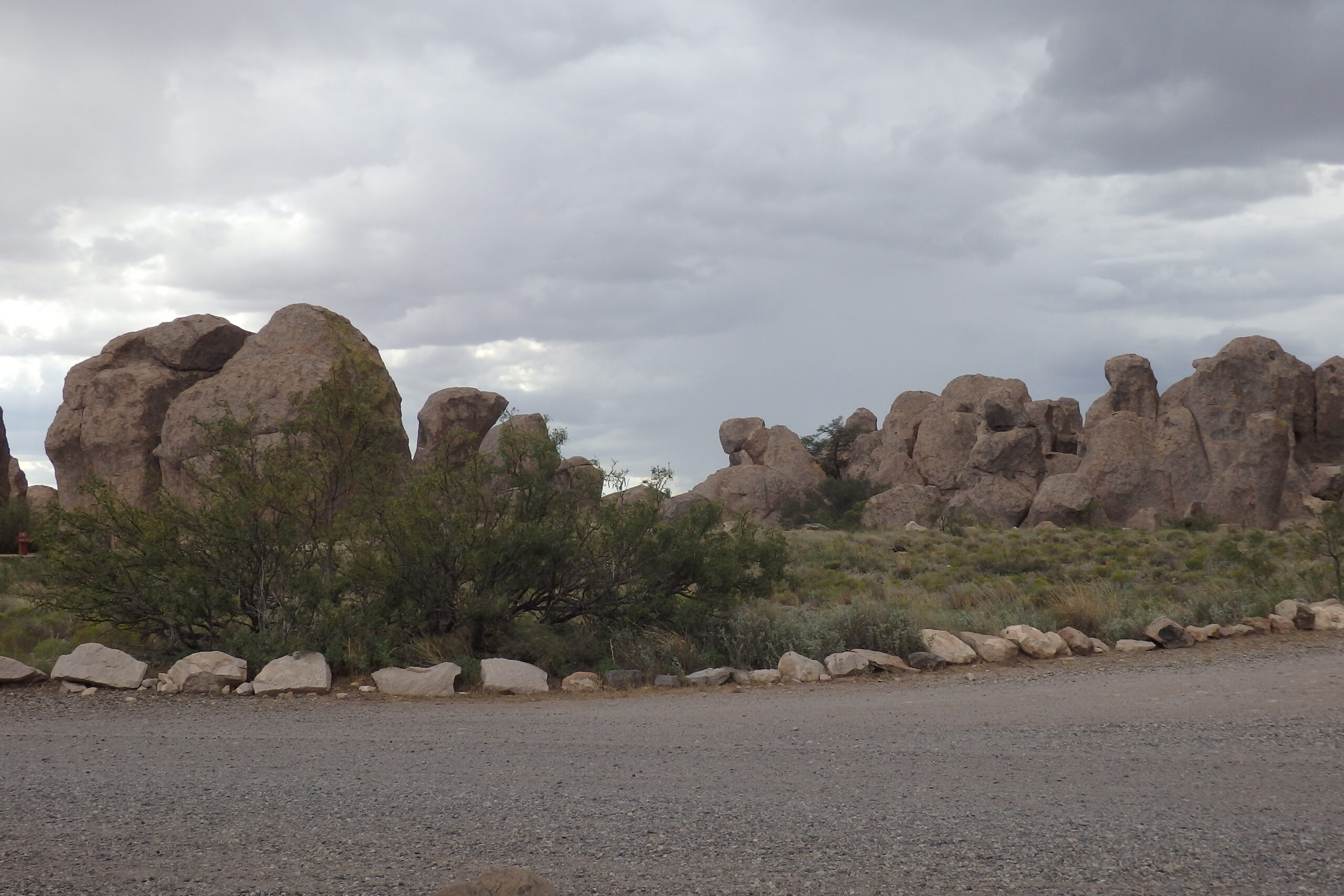 Rock formations rise dramatically under a cloudy sky. Desert vegetation and gravel paths suggest a natural park setting, inviting exploration.
