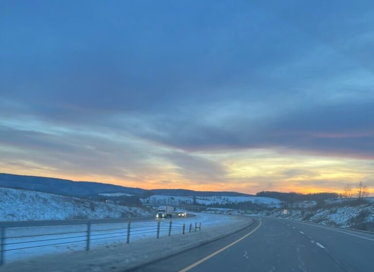 Sunset over a snowy highway, viewed from a vehicle's dashboard. The scene captures tranquil winter scenery and reflects a journey's end.