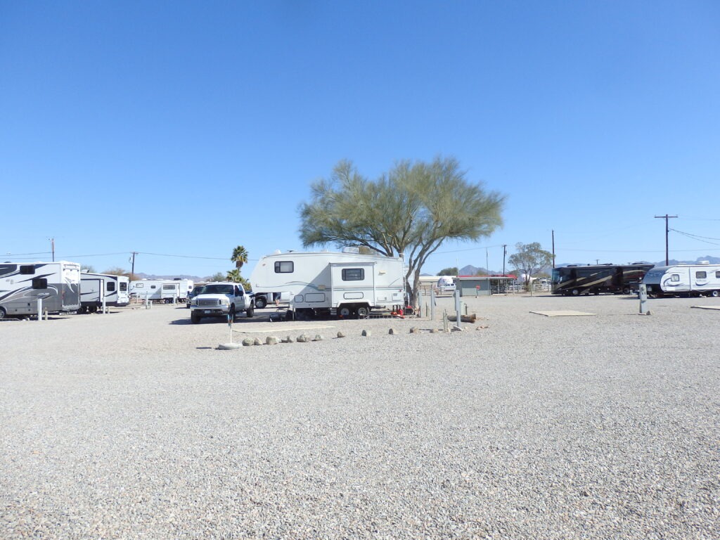 RV park featuring several parked trailers and a truck under a clear blue sky, with gravel ground and sparse vegetation, ideal for travelers.