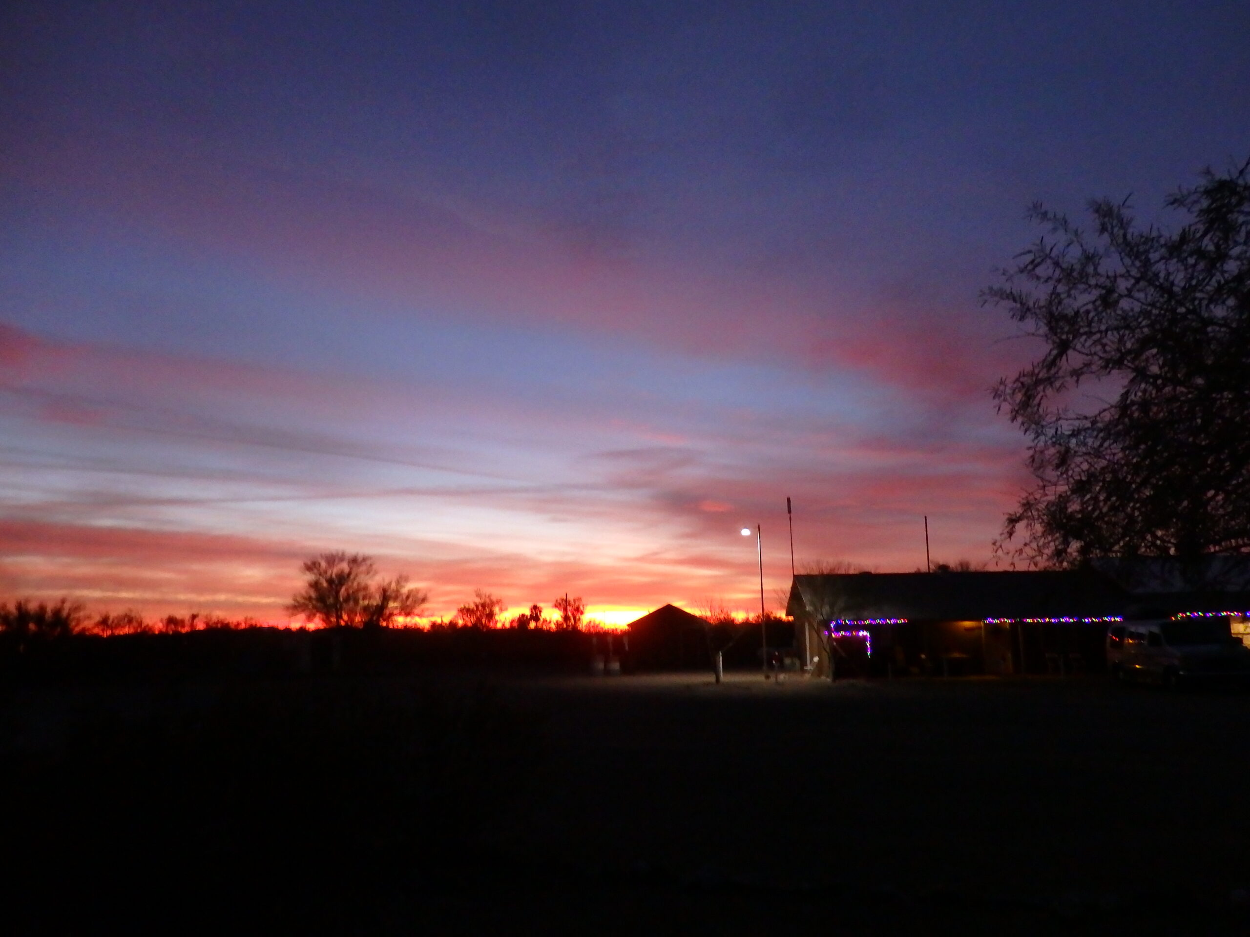 Vibrant sunset paints the sky in shades of pink and purple, silhouetting a small building decorated with lights against the desert landscape.