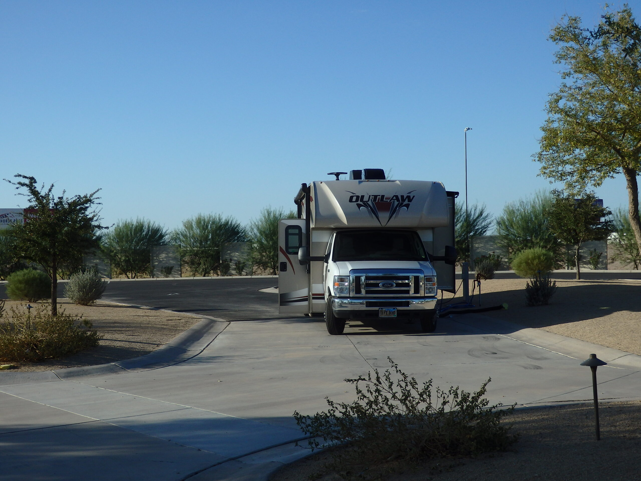 A white RV with the word "Outlaw" parked on a paved lot surrounded by desert shrubs and trees under a clear blue sky, conveying a sense of travel and adventure.