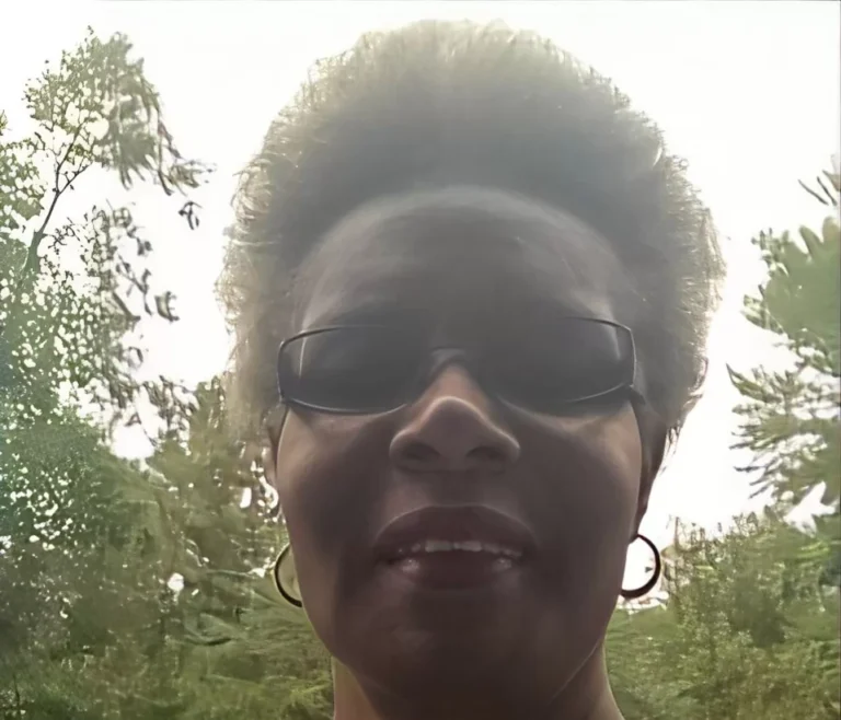 A woman with short, curly hair and sunglasses smiles for a selfie amidst a lush, green backdrop of trees, capturing a cheerful outdoor moment.