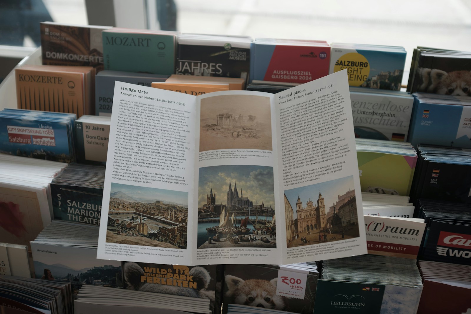 Brochure titled "Heilige Orte" highlights sacred places illustrated by Hubert Sattler, displayed among various tourist literature in Salzburg.